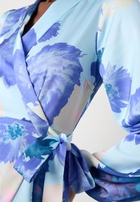 Robe portefeuille bleu clair avec de grands motifs floraux bleus et violets et un nœud noué à la taille, portée par une personne du cou à la hanche.