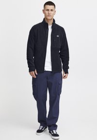 Svart fleece-zip-jakke med høy krage, med en brystlomme. Kombinert med marineblå cargo-bukser og svarte joggesko.