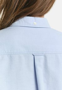 Lunghi capelli castani su una camicia colletto azzurro chiaro che supera la spalla, con pince sul retro e piccolo dettaglio a bottone bianco.