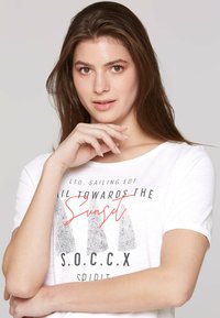 Camiseta blanca de manga corta con un estampado gráfico que incluye formas triangulares y el texto "Navega Hacia el Atardecer" en negro y rojo.
