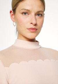 Vestido rosa claro com gola alta e painel superior transparente com bordas em forma de onda; textura suave; modelo usando pequenos brincos de argola circulares.