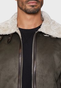 Veste marron doublée de peau de mouton avec une fermeture éclair à l'avant, extérieur en daim vert foncé, accents en cuir et fermetures à pression sur le col. Pull noir en dessous.
