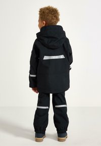 Černá nepromokavá bunda s kapucí, s reflexními pruhy na rukávech a zádech, v kombinaci s volnými černými kalhotami a gumovou obuví.