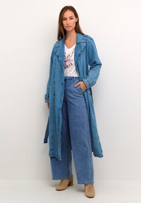 Trench coat in denim di colore azzurro chiaro, con vita regolabile, maniche ampie e grandi bottoni, abbinato a jeans a gamba larga e una maglietta con stampa floreale.