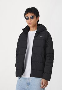 Svart puffjacka med huva, quiltad design och ett litet märke på bröstet. Bärs över en vit skjorta och kombineras med ljusa denimjeans.