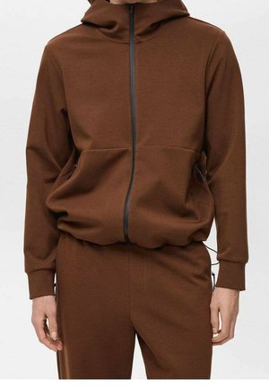 Man draagt een bruine hoodie met rits en bijpassende bruine broek, handen ontspannen langs zijn zij tegen een witte achtergrond.