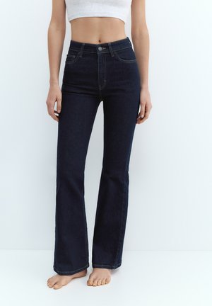 Kobieta ubrana w ciemnoniebieskie dzwony z wysokim stanem i biały krótki top, stojąca boso na jednolitym białym tle.