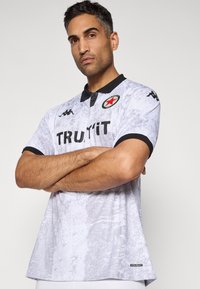 Homme avec les bras croisés portant un polo blanc texturé avec un col noir, des bords de manches noirs, des logos et le texte "TRUYYiT".