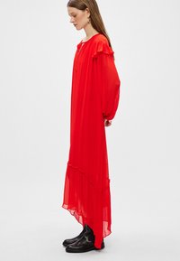 Vestido de chiffon rojo con acentos de volantes, mangas largas y una silueta fluida. Presenta un dobladillo asimétrico y se lleva con botas negras hasta el tobillo.