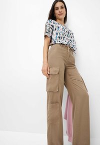 Beige cargobroek met grote zijzakken en riemlussen, gecombineerd met een gedrukt blouse met korte mouwen in pastelkleuren met bladdesigns.