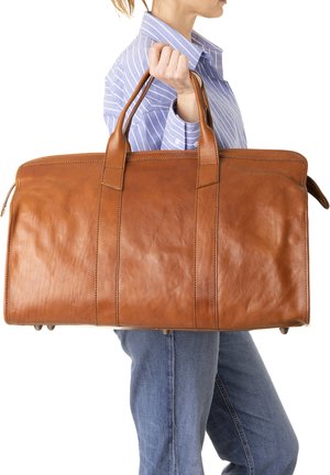 Persoon die een grote, bruine leren duffeltas bij de handvatten draagt, gekleed in een blauw gestreept overhemd en blauwe spijkerbroek, zijaanzicht tegen een witte achtergrond.