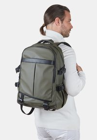 Khaki zelena nahrbtnik s strukturiranim dizajnom, ki vsebuje črn poudarni trak, prilagodljive naramnice in več zunanjih žepov.