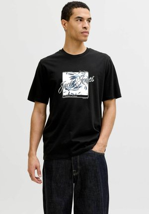 Młody mężczyzna w czarnej koszulce z napisem i wzorem "Jack & Jones", w ciemnych dżinsach, stojący z jedną ręką w kieszeni.