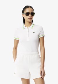 Polo bianco con colletto a righe verdi e gialle, logo ricamato sul petto, abbinato a pantaloni corti bianchi con cintura elastica. Occhiali da sole scuri indossati.