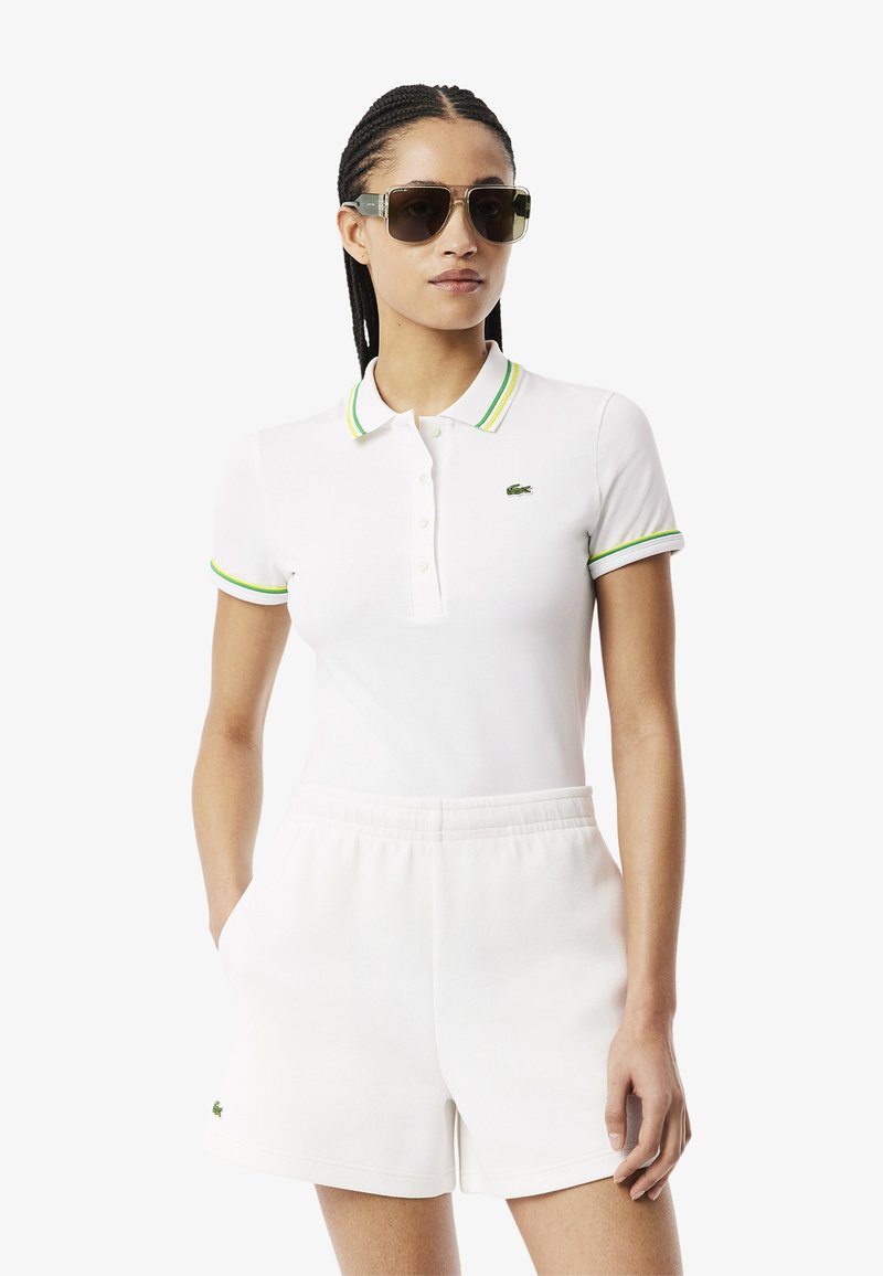 Polo bianco con colletto a righe verdi e gialle, logo ricamato sul petto, abbinato a pantaloni corti bianchi con cintura elastica. Occhiali da sole scuri indossati.