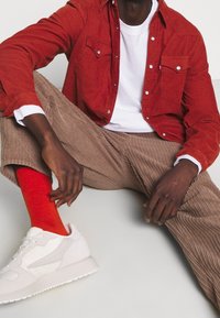 Rode corduroy blouse, wit T-shirt, bruine corduroy broek, oranje sokken en witte sneakers. Kenmerkend zijn de gestructureerde stof en contrasterende kleuren.