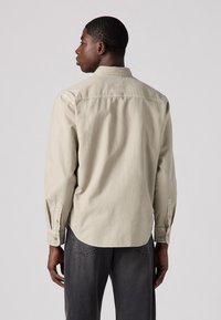 Lichtbeige langslavende blouse met een button-down kraag, voorzien van zijplooien en knoopsluitingen bij de mouwen. Gladde textuur met een ontspannen pasvorm.