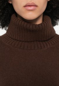 Pulover maro cu guler înalt și textură tip tricot cu dungi. Focalizat pe partea centrală, arătând detaliile cusăturilor și materialul moale.