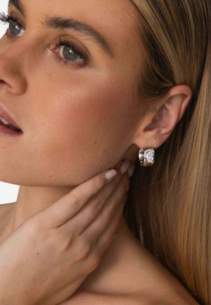 Silberne Creolen mit einem transparenten Edelsteinakzent, polierter Oberfläche und rundem Design, getragen von einer Person, die ihren Hals berührt.