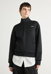 Black zip-up jacket with high collar, made of soft fabric. Features logo on chest and pockets. Worn over a white shirt.