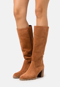 Botas de ante marrón hasta la rodilla con un diseño estrecho, cremallera lateral y tacón apilado. La superficie tiene una textura suave y detalles mínimos.