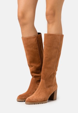 Botas de ante marrón hasta la rodilla con un diseño estrecho, cremallera lateral y tacón apilado. La superficie tiene una textura suave y detalles mínimos.