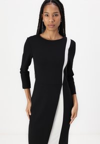 Crna haljina s tričetvrtinim rukavima, koja ima bijelu dijagonalnu prugu i prorez. Glatka, blago teksturirana tkanina. Jednostavna okrugla izrez.