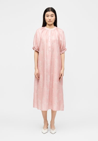 Mujer con cabello largo y oscuro que lleva un vestido midi rosa suelto con mangas abullonadas y pequeños botones, combinado con tacones blancos de punta afilada.