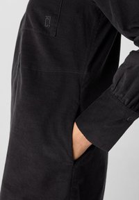 Schwarzes Cordkleid mit gerader Silhouette, seitlichen Taschen und geknöpften Manschetten mit feiner Rippstruktur. Branding auf der Rücktasche.