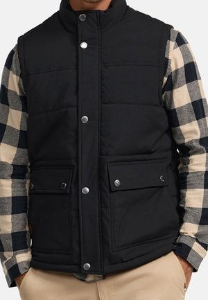 Černá prošívaná vesta na patentky nošená přes černo-béžovou kostkovanou košili s dlouhým rukávem, doplněná béžovými kalhotami, ruce v kapsách.