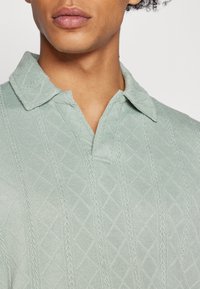 Polo de malha verde claro com um padrão de diamante texturizado e colarinho pontudo. Apresenta um delicado detalhe de malha em cabos ao longo das costuras.