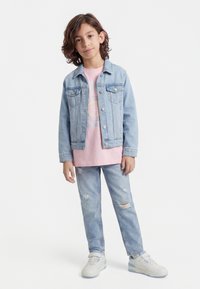 Tânăr cu păr până la umeri purtând o geacă de denim albastru deschis, tricou roz cu imprimeu, blugi rupți și teniși albi.