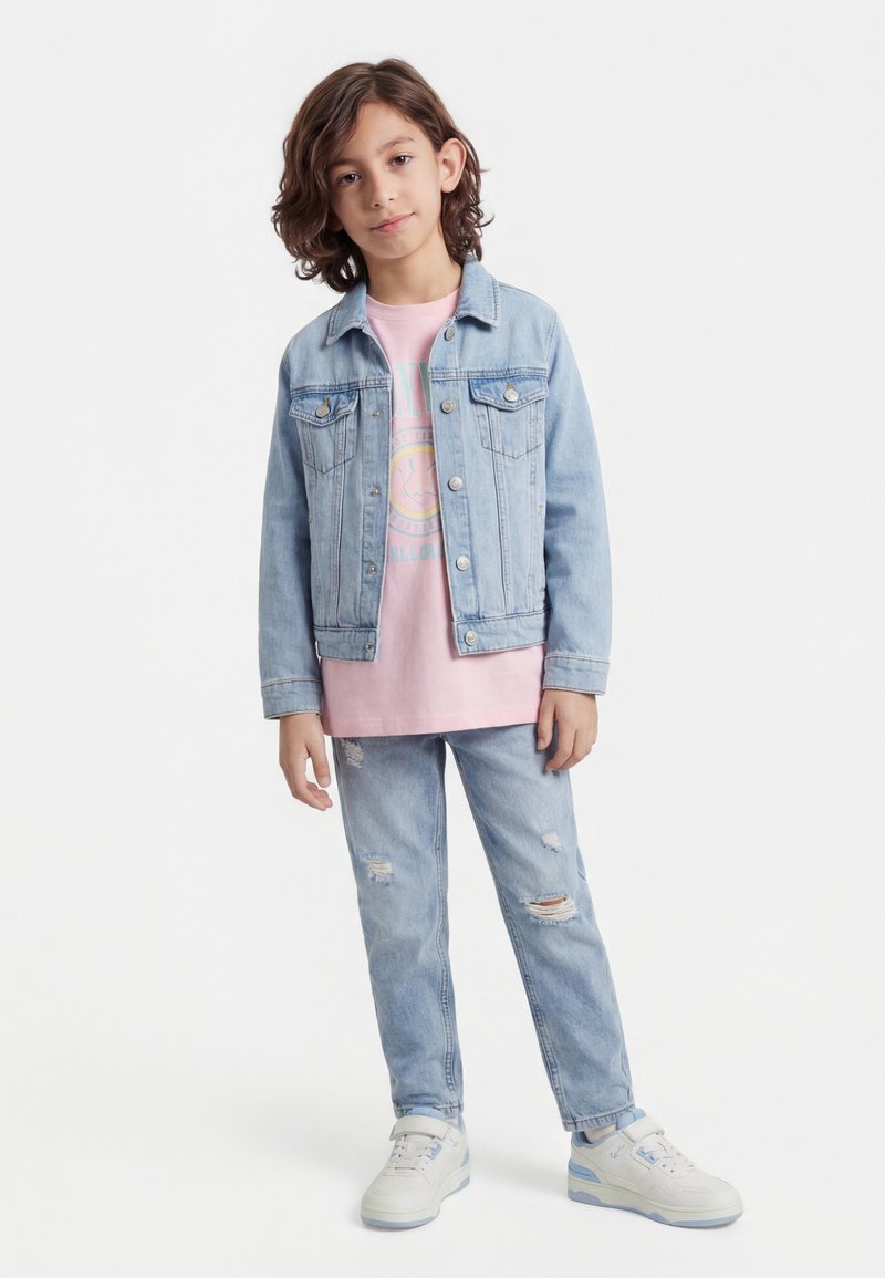 Tânăr cu păr până la umeri purtând o geacă de denim albastru deschis, tricou roz cu imprimeu, blugi rupți și teniși albi.