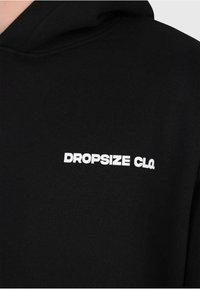 Schwarzer Kapuzenpullover mit weißem Schriftzug "DROPSIZE CLO." auf der oberen Brust, zeigt einen Teil des Halsausschnitts und des Schulterbereichs.
