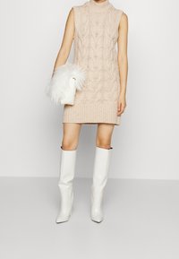 Abito senza maniche lavorato a maglia di colore rosa chiaro con motivo a treccia testurizzato; abbinato a una borsa a tracolla in pelliccia ecologica bianca e stivali alti fino al ginocchio bianchi e lucidi.