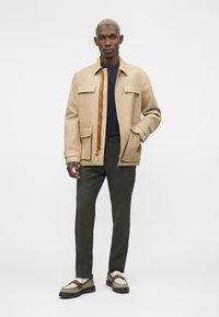 Homme portant une veste beige avec de grandes poches, une chemise noire, un pantalon foncé et des mocassins beige et marron, debout devant un fond uni.
