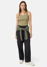 Olivegrünes Tanktop, lockere schwarze Hose, graue Strickjacke um die Taille gebunden und weiße Sneakers mit Gummisohlen.