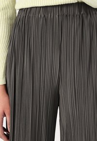 Ingrandimento di una persona che indossa pantaloni plissettati grigio scuro e un maglione a costine giallo chiaro, con la mano appoggiata accanto ai pantaloni.