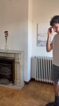 Mand, der justerer sine briller nær en hvid radiator, med en marmorpejs, der holder en vase med røde blomster, og et maleri på væggen i baggrunden.