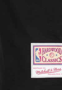 La camiseta de tela negra presenta una etiqueta blanca rectangular con el logo multicolor de la NBA y el texto "HARDWOOD CLASSICS". Incluye la marca "Mitchell & Ness".