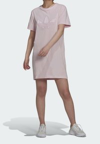 Růžové bavlněné šaty ve stylu trička s krátkými rukávy, s tónovým logem Adidas na hrudi a rovnym lemem. Nosí se s bílými sportovními botami.