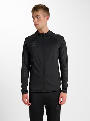 Jeune homme portant une veste de sport noire zippée et un pantalon assorti, regardant vers la gauche sur un fond blanc uni.