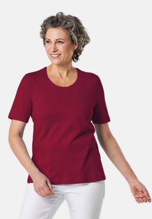 T-shirt rouge à manches courtes avec un col rond, fabriqué en tissu doux. Le modèle le porte avec un pantalon blanc, mettant en valeur une coupe décontractée.