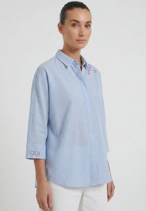 Femme aux cheveux plaqués en arrière portant une chemise boutonnée à fines rayures bleu et blanc avec « oui » brodé sur le poignet et un pantalon blanc.