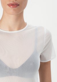 Femme portant un haut blanc transparent à manches courtes par-dessus un soutien-gorge noir, montrant des lèvres avec un rouge à lèvres nude brillant ainsi qu'une partie du cou et des épaules.