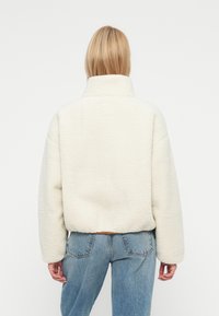 Fleecasta jakna u boji slonovače s visokim ovratnikom, spuštenim ramenima i teksturiranim završetkom. Uparen s svijetloplavim trapericama.