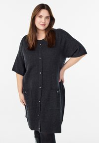 Tamsiai pilkas kardiganas su trumpomis rankovėmis, sagų užsegimu ir dviem priekinėmis kišenėmis. Pagamintas iš minkštos, tekstūruotos medžiagos. Minimalistinio dizaino su sidabriniais akcentais.