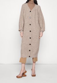 Kvinna klädd i en lång beiga knäppt kofta över en brun polotröja och korta byxor, tillsammans med trälädersandaler med genomskinliga remmar.