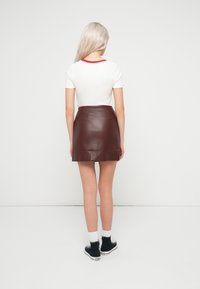 Λευκό πουκάμισο με κόκκινο γιακά, συνδυασμένο με μίνι φούστα από καφέ δέρμα. Το μοντέλο φορά μαύρα αθλητικά παπούτσια και λευκές κάλτσες. Απλή φόντο.