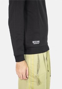 Černá mikina s dlouhým rukávem s žebrovanými manžetami a lemem, s bílým logem "Moschino Underwear" na levé straně, doplněná světle zelenými kalhotami.